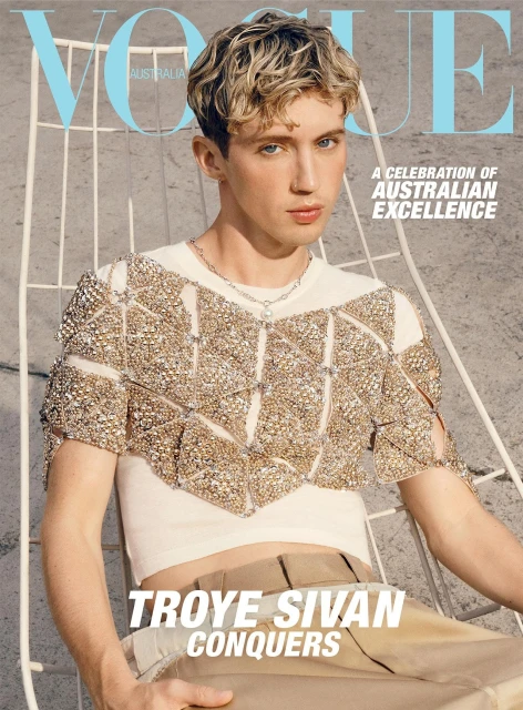 Vogue Australia cover featuring Troye Sivan in a white and gold beaded top, tan pants, and blond hair, set against a grey backdrop.