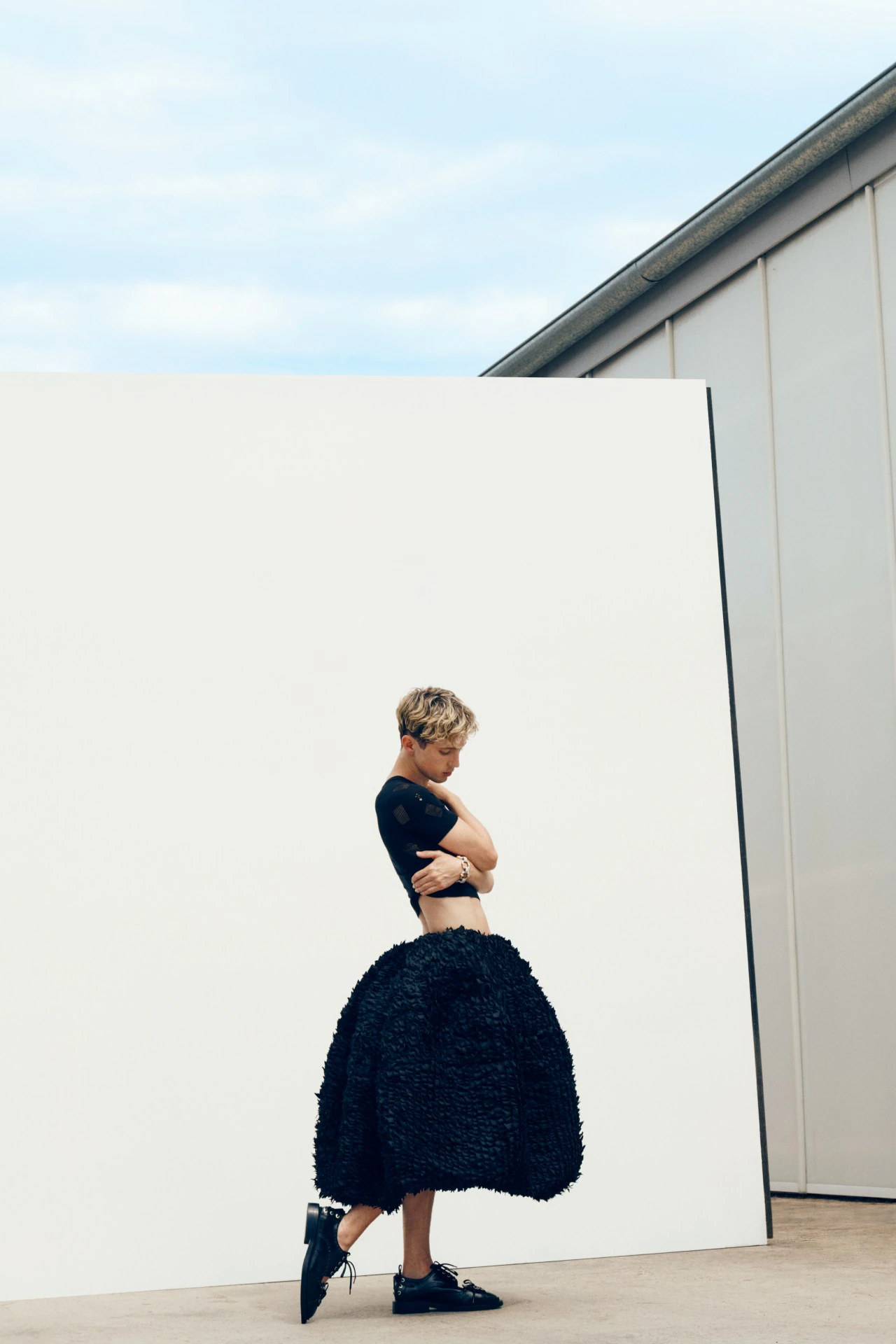 Fashion shot of a person with short hair posing in a black crop top and voluminous black skirt, standing in front of a white wall.