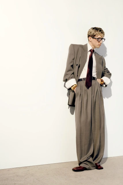 A man in a grey double-breasted suit, white shirt, and maroon tie poses against a white wall in a studio environment.