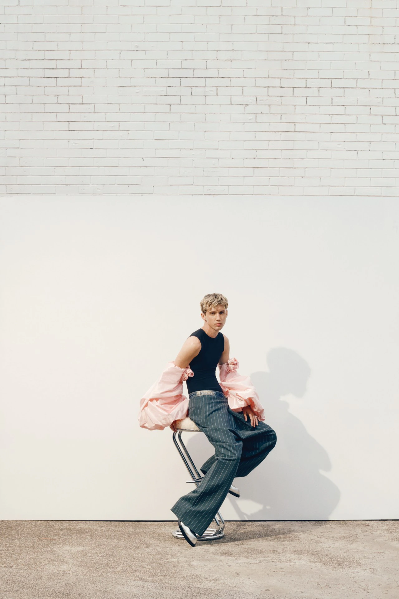 A young adult male model sits on a stool wearing a black tank top, grey pinstripe pants, and a pink sleeve bolero in a studio.