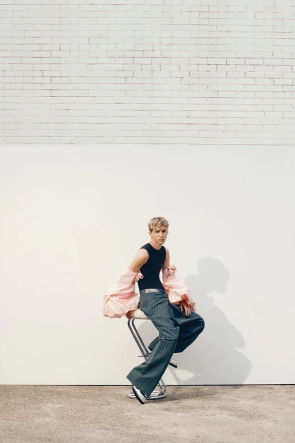A young adult male model sits on a stool wearing a black tank top, grey pinstripe pants, and a pink sleeve bolero in a studio.