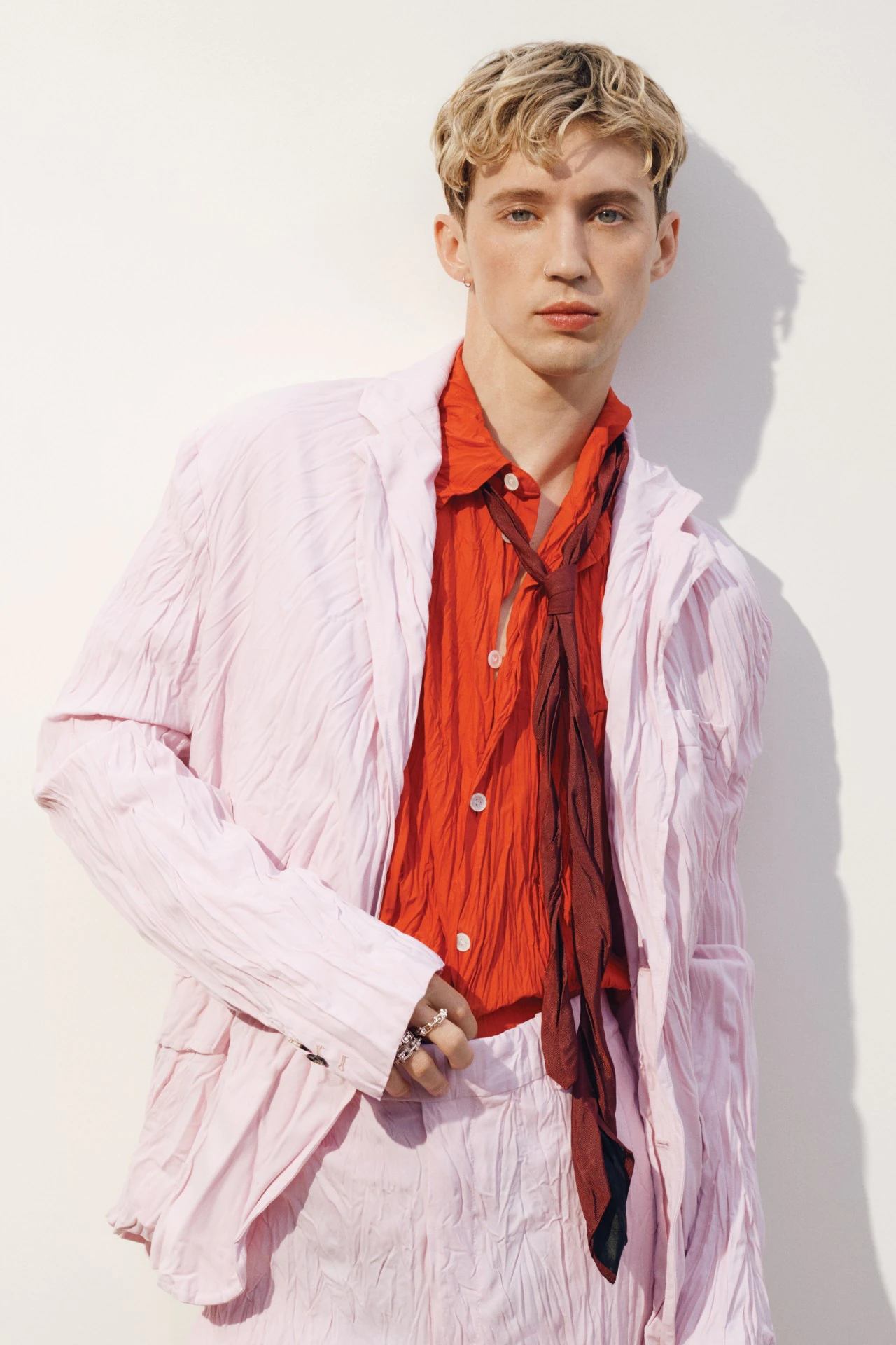 Young male model with blond hair poses in a light pink suit with a red button-down shirt and loosely-tied brown tie against a white wall.