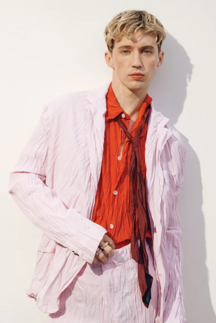 Young male model with blond hair poses in a light pink suit with a red button-down shirt and loosely-tied brown tie against a white wall.