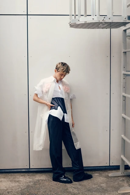 Woman in sheer coat, white shirt, and pinstripe pants poses indoors, leaning against a wall with metal accents and a ladder.