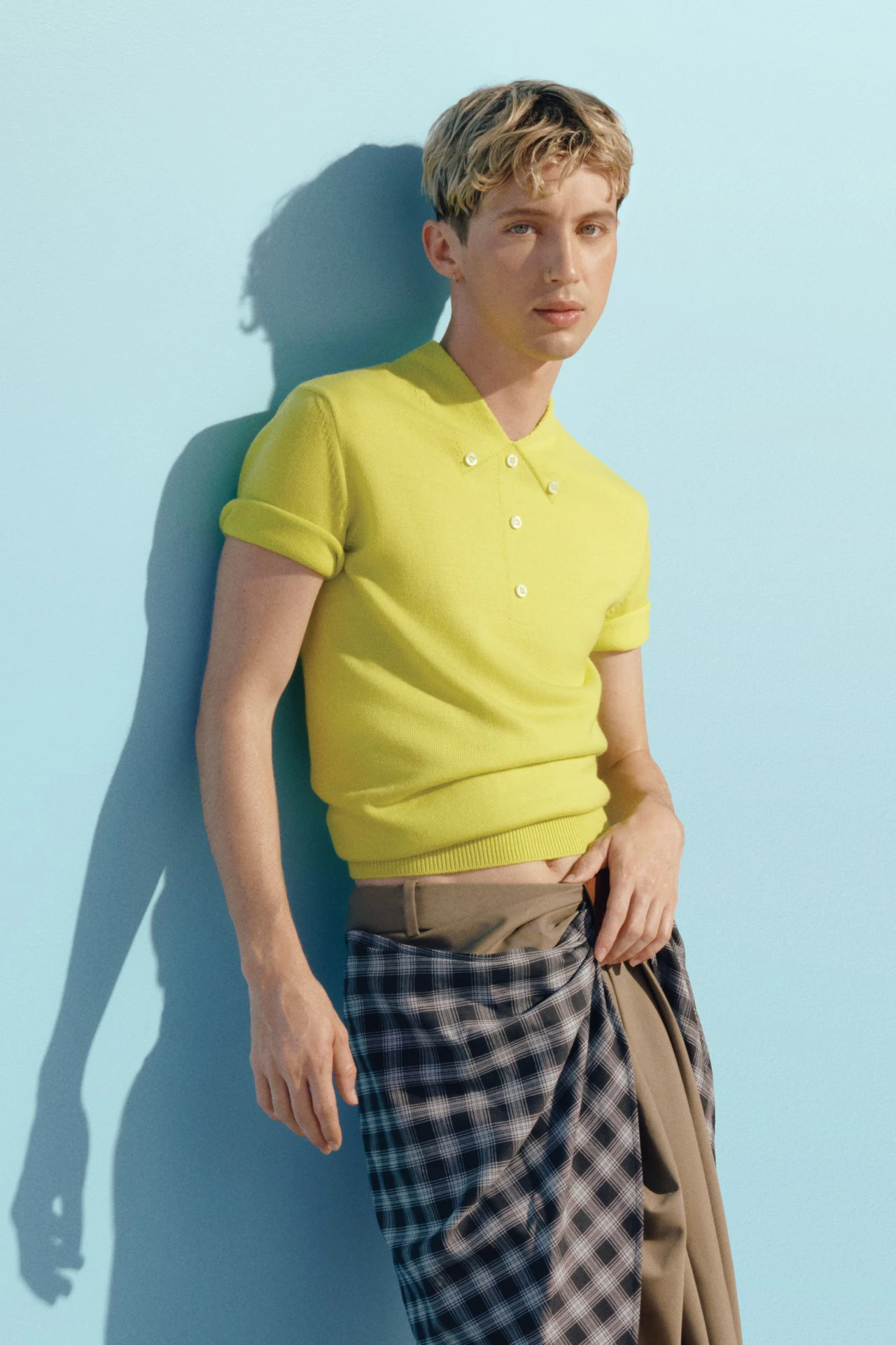Young adult in stylish portrait with bright yellow polo shirt and plaid skirt, against a light blue backdrop with studio lighting.