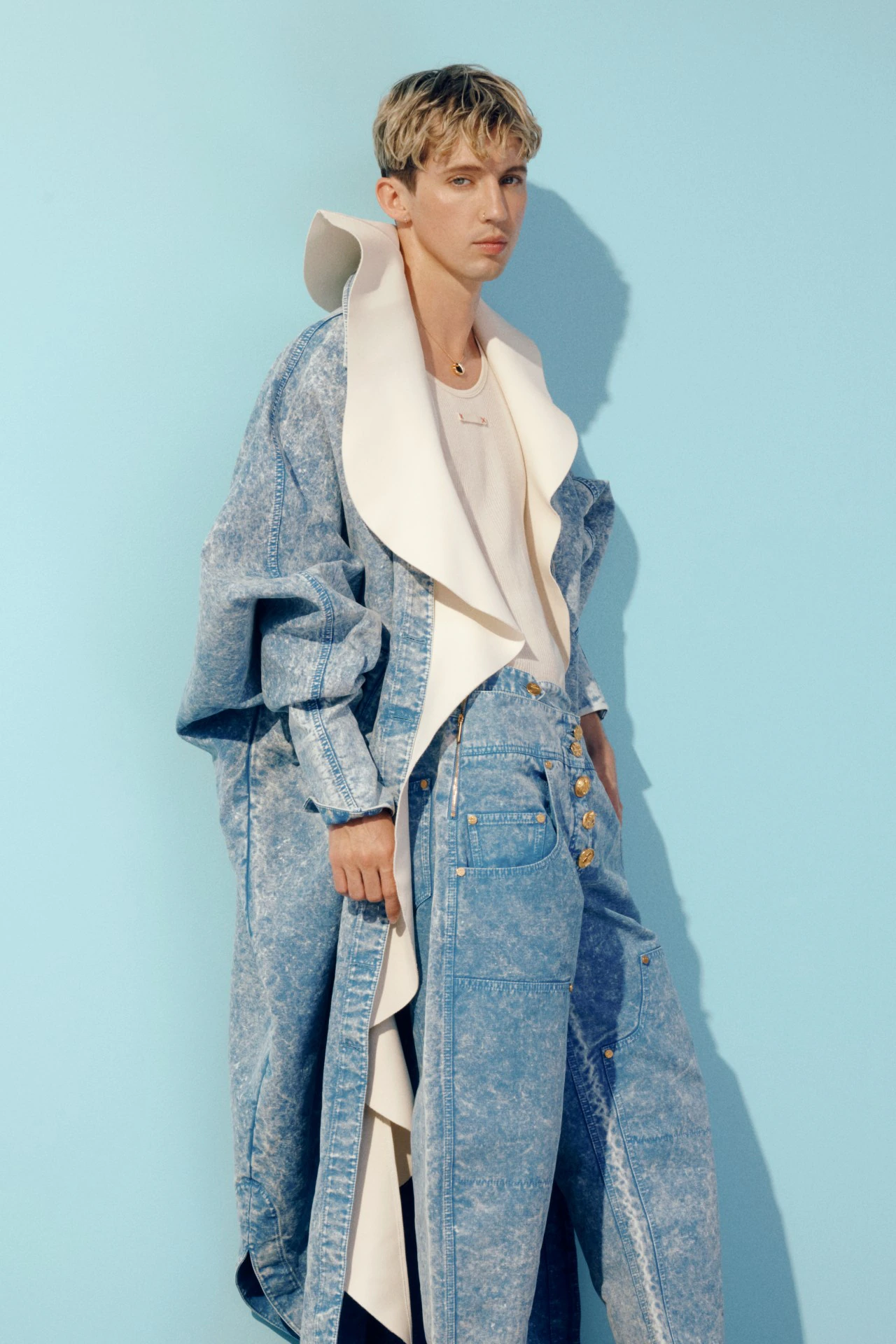Young adult stands in a fashion pose wearing an acid-wash denim coat and pants against a light blue backdrop in a studio setting.