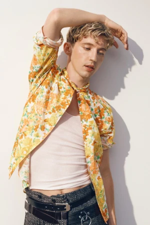 Portrait of a young adult male model wearing a floral shirt and black belt against a white background, striking a casual pose with hand raised.