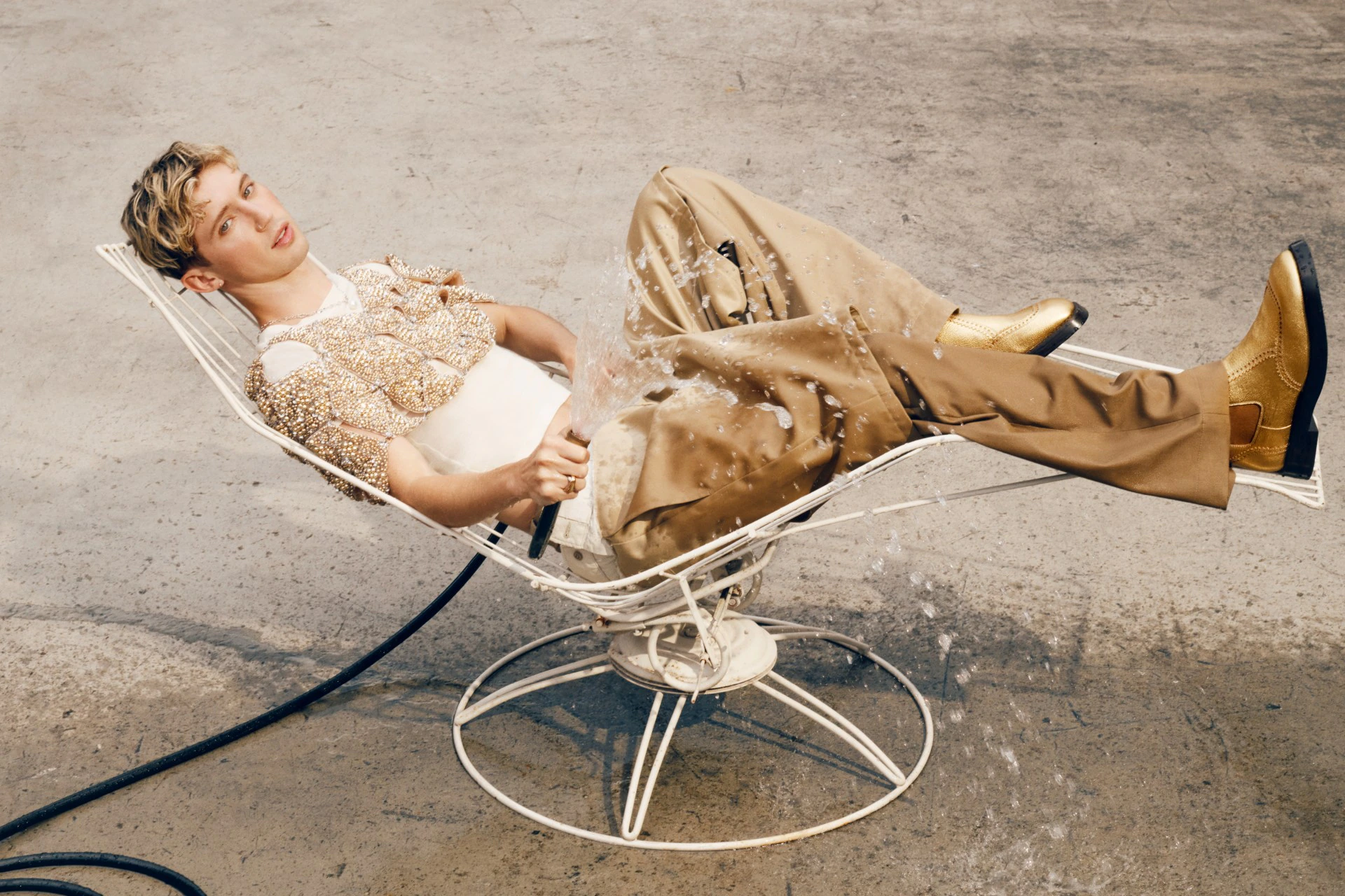 Man in tan outfit reclines on white wire chair, is sprayed with water in a light outdoor setting with gold boots. 