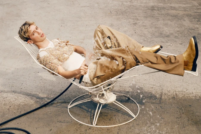 Man in tan outfit reclines on white wire chair, is sprayed with water in a light outdoor setting with gold boots. 