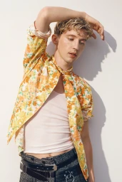 Troye Sivan posing in a yellow floral patterned shirt and textured skirt against a plain white studio background.