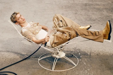 Troye Sivan reclining in a white wire chair while holding a water hose against a concrete background.