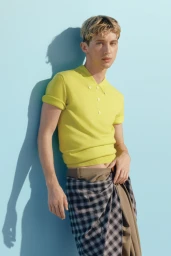 Troye Sivan in a yellow polo shirt and plaid skirt standing against a light blue studio wall for Vogue Australia.
