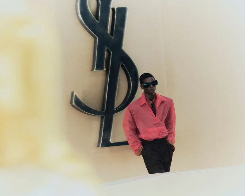 An adult male in a salmon-colored shirt and dark pants poses next to a large, black YSL logo against a beige wall in a studio setting.