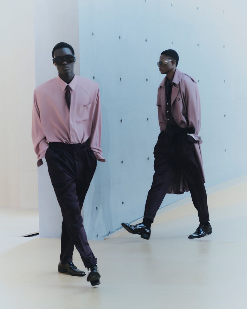 Two dark-skinned adult males styled in mauve dress shirts, ties, and dark trousers pose indoors against a patterned pale blue wall and floor.