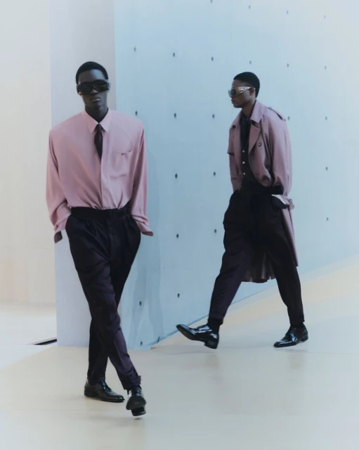 Two dark-skinned adult males styled in mauve dress shirts, ties, and dark trousers pose indoors against a patterned pale blue wall and floor.