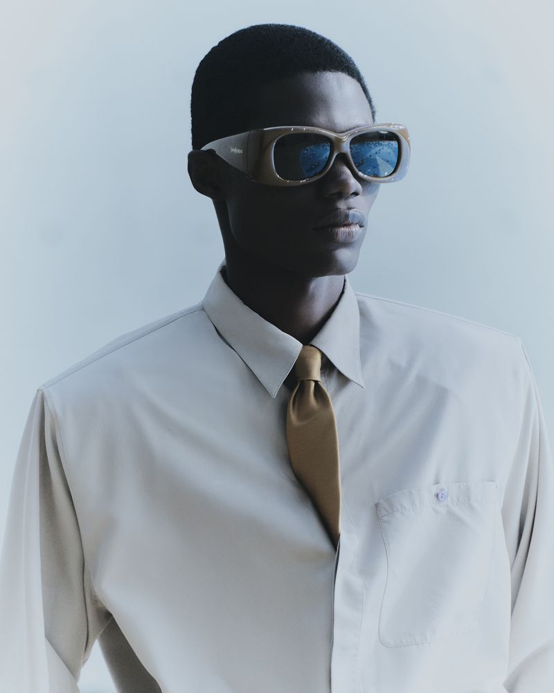 Dark-skinned adult man wearing sunglasses and a light beige button-down shirt with a brown tie poses in front of a pale blue background.