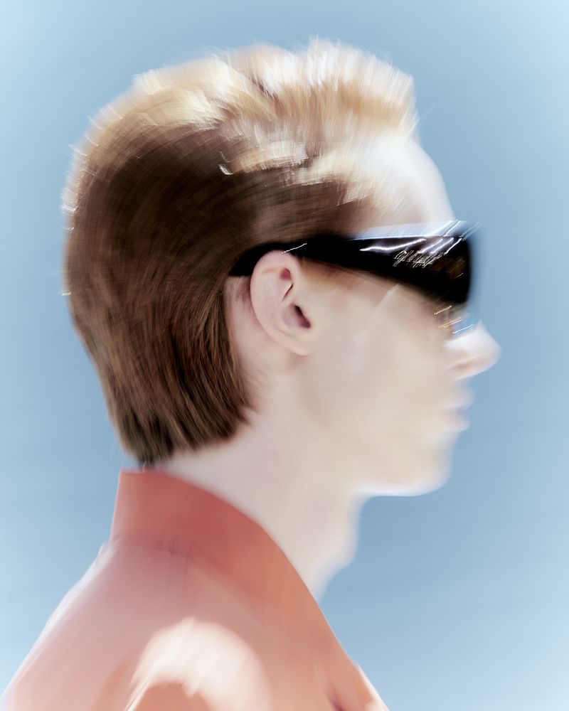 Side profile of a person with light brown hair wearing sunglasses, against a light blue background, captured with motion blur.