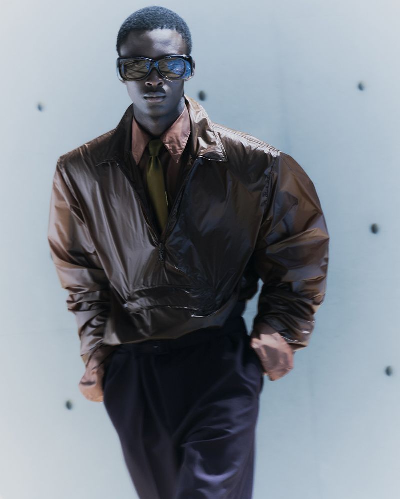 An adult male stands indoors wearing sunglasses, a brown jacket, a tan shirt, and dark pants in a fashion-editorial style portrait.