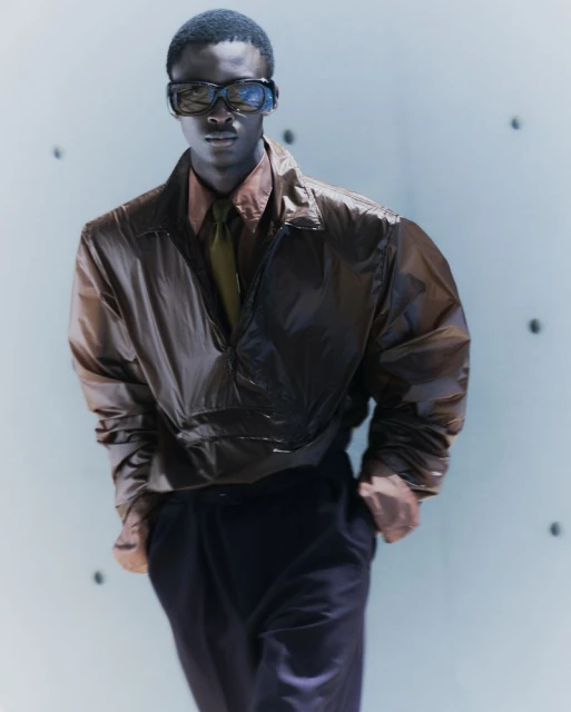 An adult male stands indoors wearing sunglasses, a brown jacket, a tan shirt, and dark pants in a fashion-editorial style portrait.