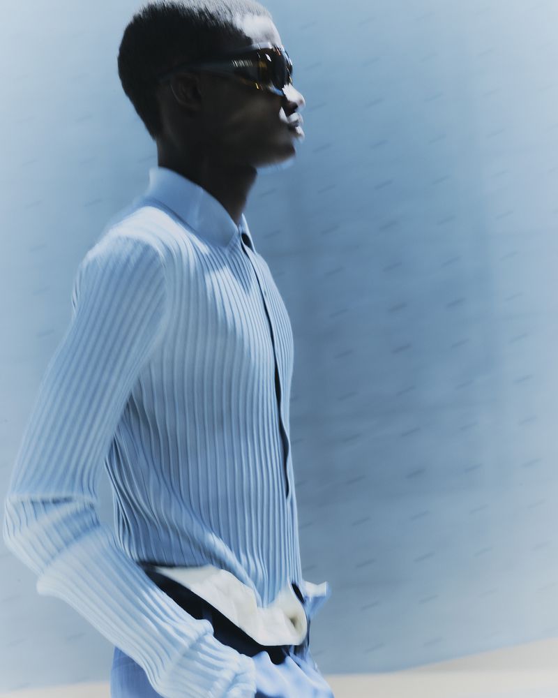 A light-skinned man in a light-blue ribbed shirt and sunglasses poses in a studio with a blurred-light blue backdrop and neutral-colored floor.