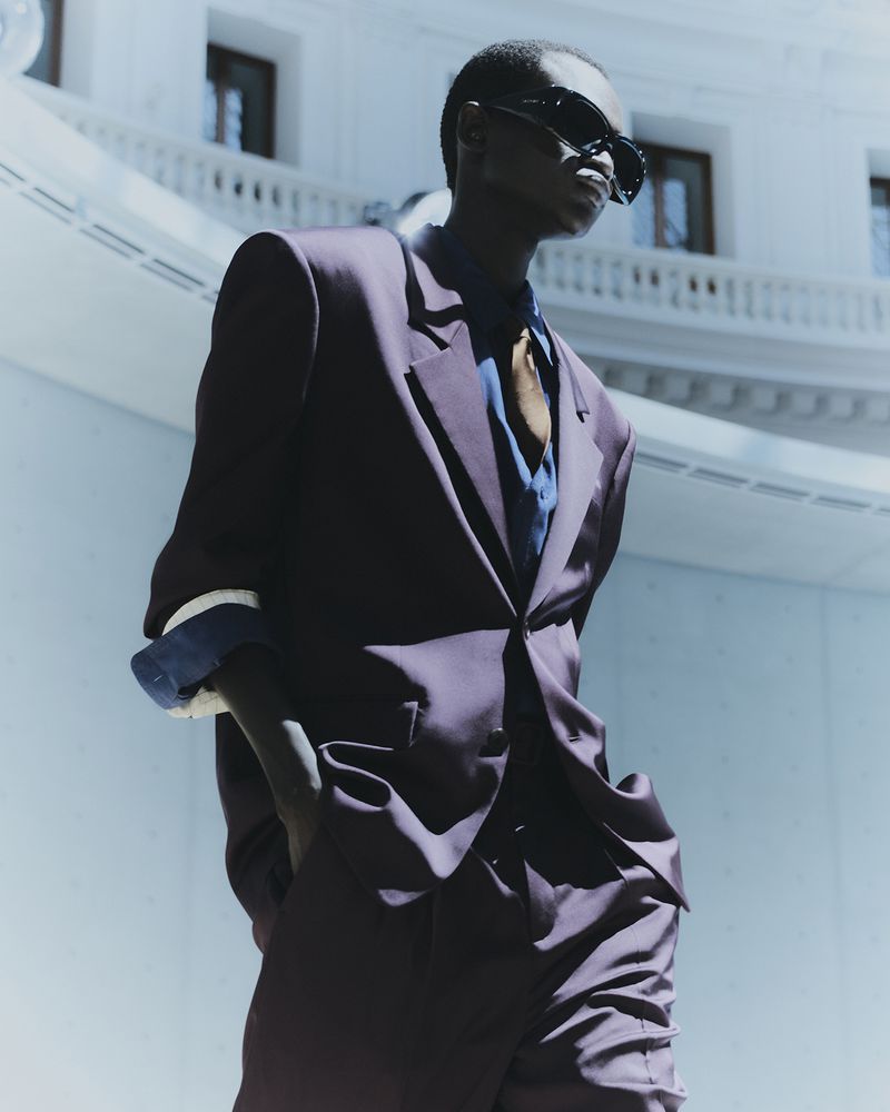 Man in a dark suit and sunglasses standing in an outdoor location with a white building and columns in the background on a sunny day.