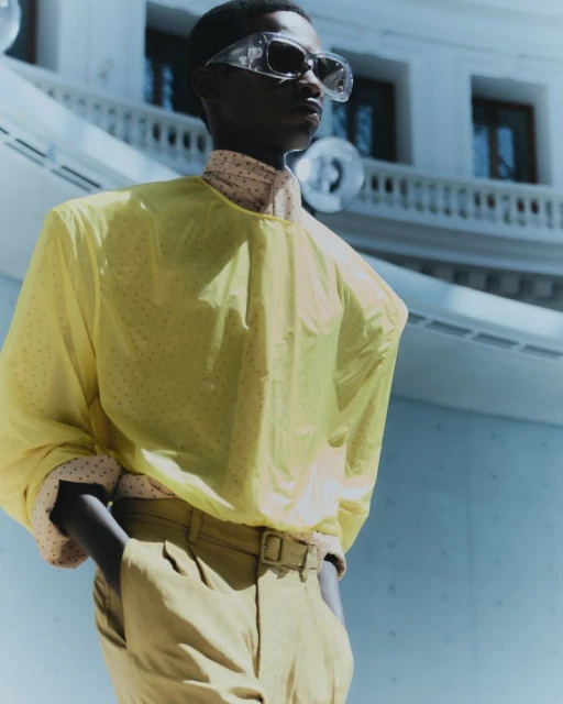 Fashionable adult male in yellow top and beige pants poses outdoors with architectural backdrop in editorial fashion shoot.