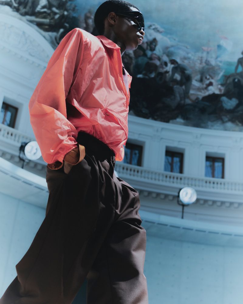 African American male model strides confidently in a salmon jacket and oversized brown trousers against a backdrop of architectural details.
