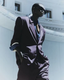 Black male model wearing an oversized purple tailored suit and dark sunglasses against a curved architectural backdrop.