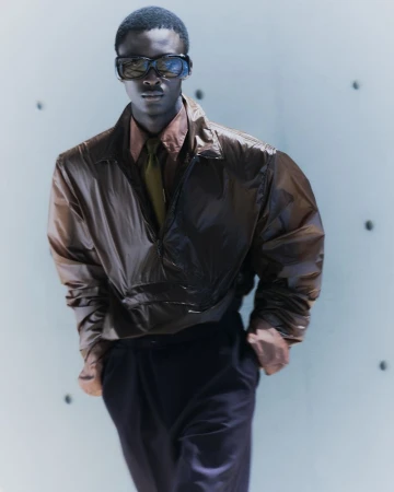 Black male model in dark sunglasses wearing an oversized bronze half-zip jacket and tie against a pale architectural wall.