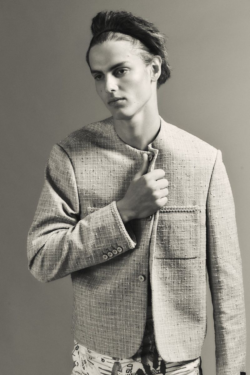 Model Esteban Monbaliu wearing a textured collarless tweed jacket and black headband against a grey background.