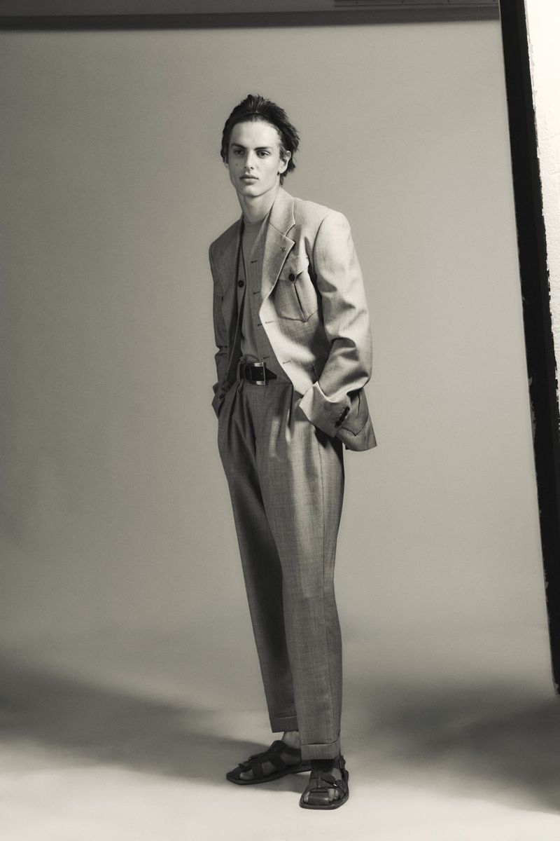 Esteban Monbaliu poses in a tailored grey suit with pleated trousers and leather sandals against a studio backdrop.