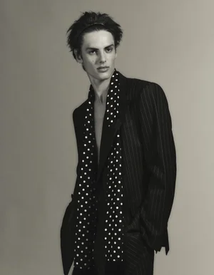 Esteban Monbaliu wearing a pinstripe blazer and a black and white polka dot scarf against a grey studio backdrop.