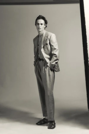 Esteban Monbaliu poses in a tailored grey suit with pleated trousers and leather sandals against a studio backdrop.