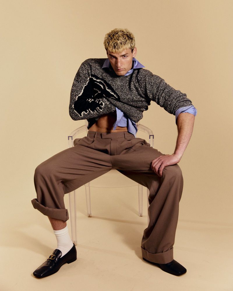 Model with bleached hair seated on a clear chair in a grey knit sweater and brown wide-leg pleated trousers on a beige backdrop.