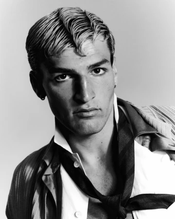 Black and white close up portrait of a young man with styled wavy hair wearing an unbuttoned shirt and loose tie.