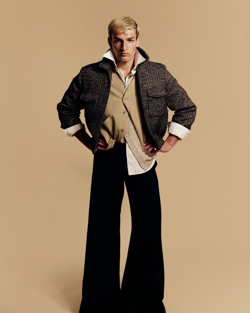 Model in flared black trousers and a textured cropped jacket posing with hands on hips against a beige studio backdrop.
