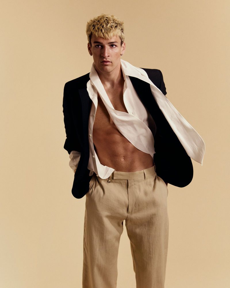 Male model with bleached blonde hair wearing an open black blazer, unbuttoned white shirt, and beige trousers on a tan backdrop.