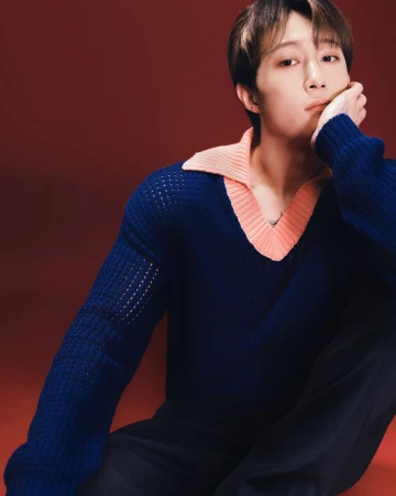 An East Asian man seated against a dark red backdrop wearing a blue knit sweater with a peach collar.