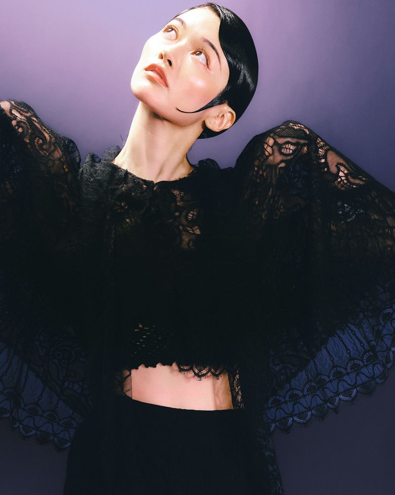East Asian woman with sleek sculpted hair looking upward while wearing an intricate sheer black lace top against a violet studio background.