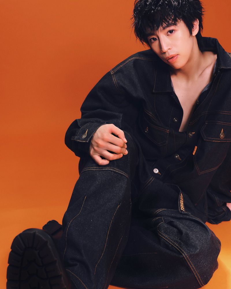 East Asian man in an oversized black denim jacket and wide-leg trousers seated on the floor against a solid orange background.