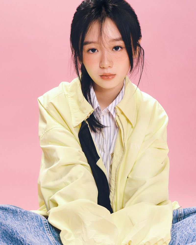 East Asian young woman in a pale yellow jacket and blue jeans sitting against a solid pastel pink background.