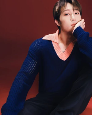 An East Asian man seated against a dark red backdrop wearing a blue knit sweater with a peach collar.