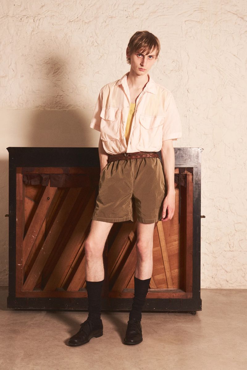 Model standing in front of an upright piano wearing a light pink button-down shirt, brown shorts, and black socks with shoes.