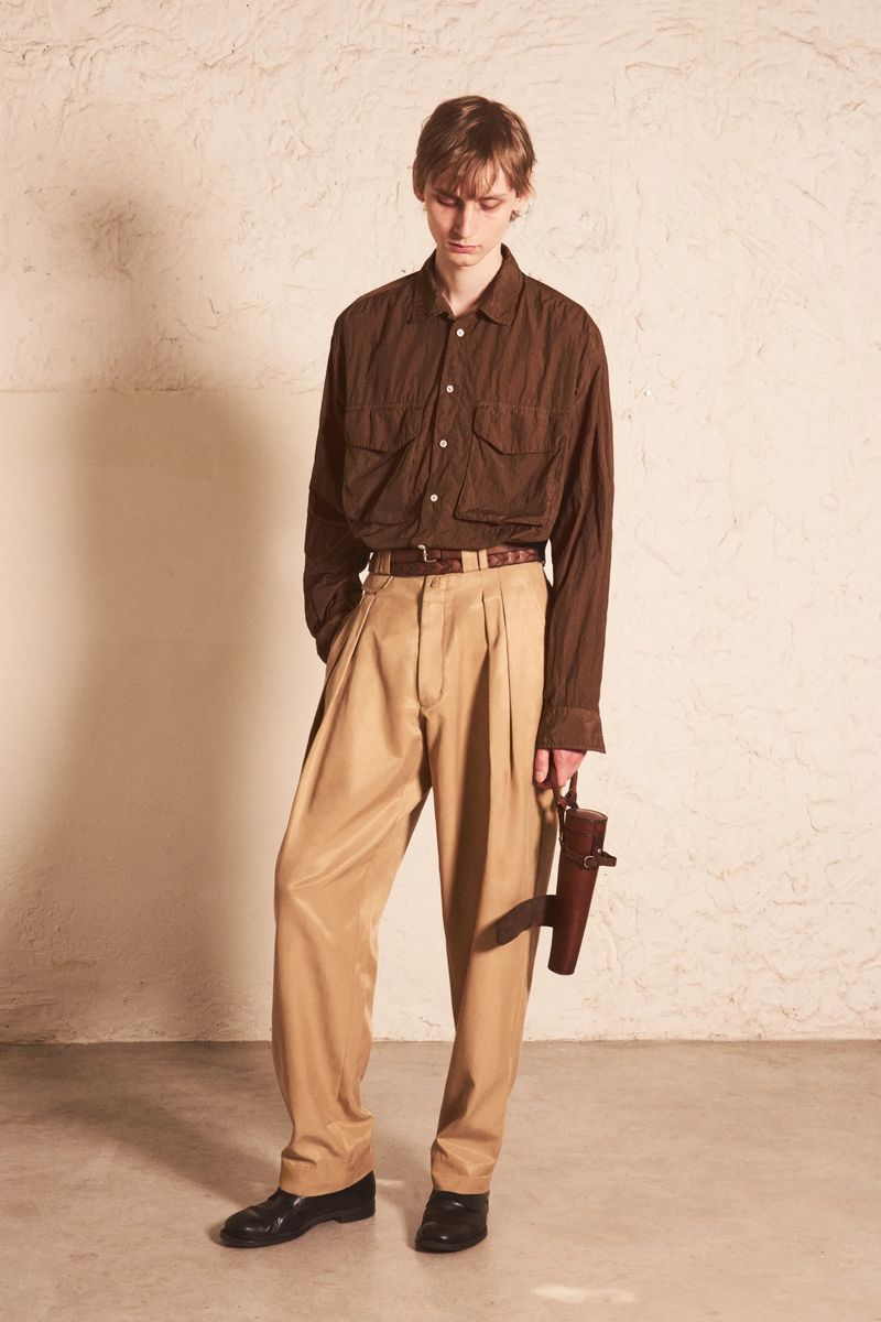 Male model in a brown button-up utility shirt and pleated beige trousers standing against a textured plaster wall.
