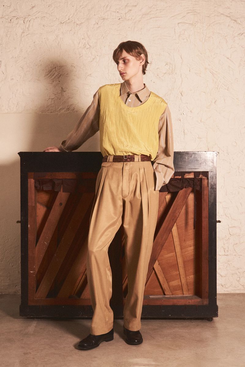 Model leaning against a dark upright piano wearing a yellow textured vest, beige pleated trousers, and a collared shirt.