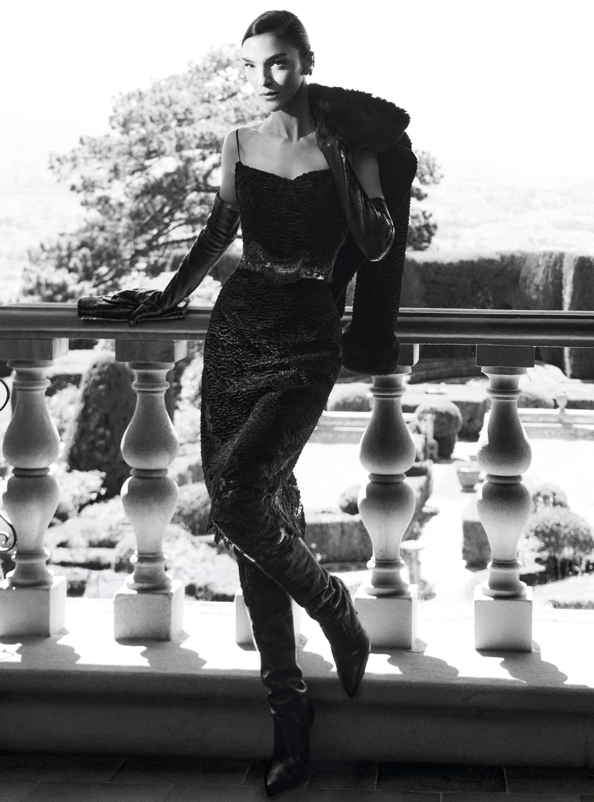 Mariacarla Boscono in a black dress, leather gloves, and boots standing on a stone balcony with a balustrade for Ermanno Scervino.