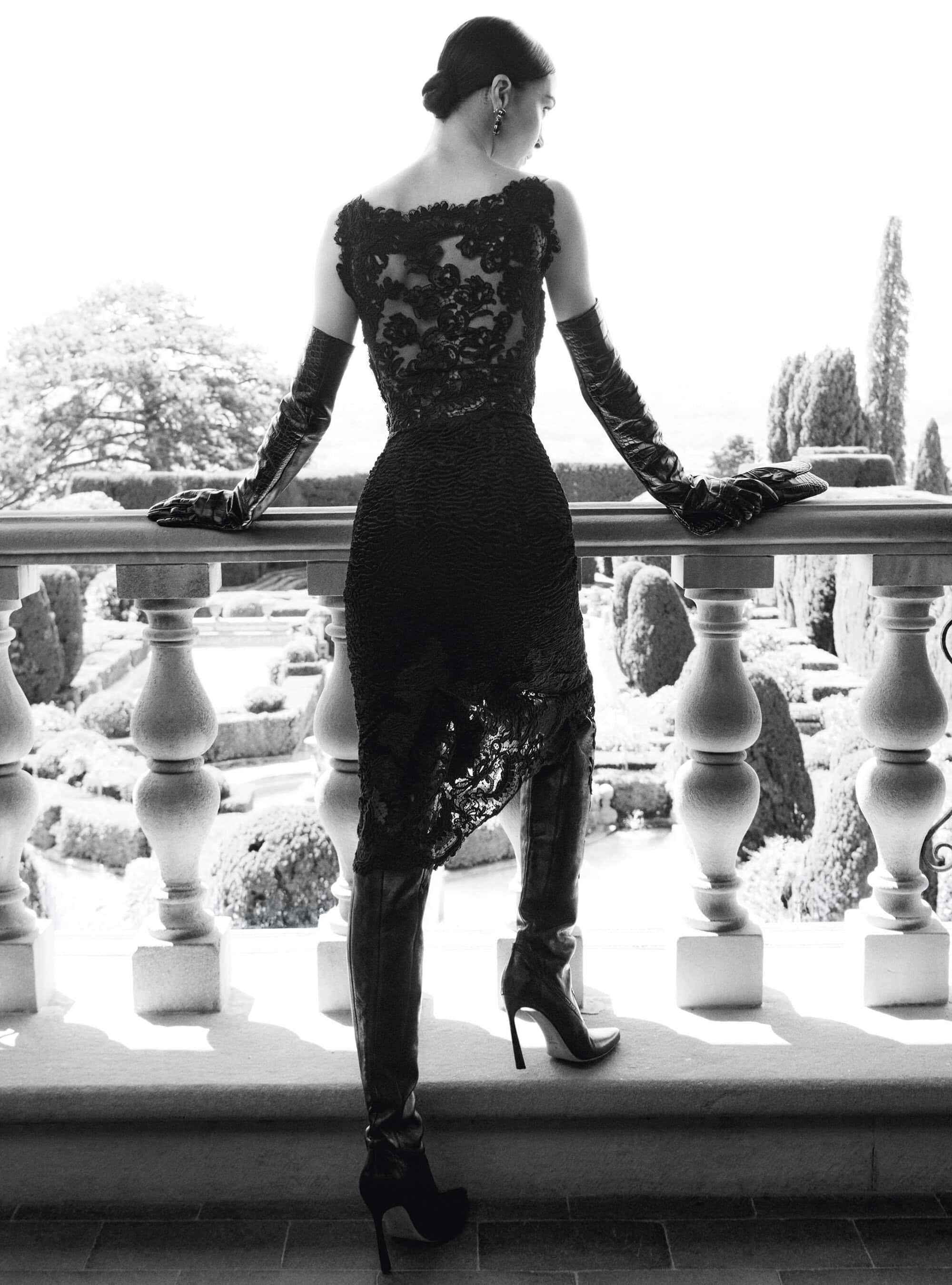 Mariacarla Boscono seen from behind wearing a black lace dress, long leather gloves, and heeled boots on a stone balustrade.