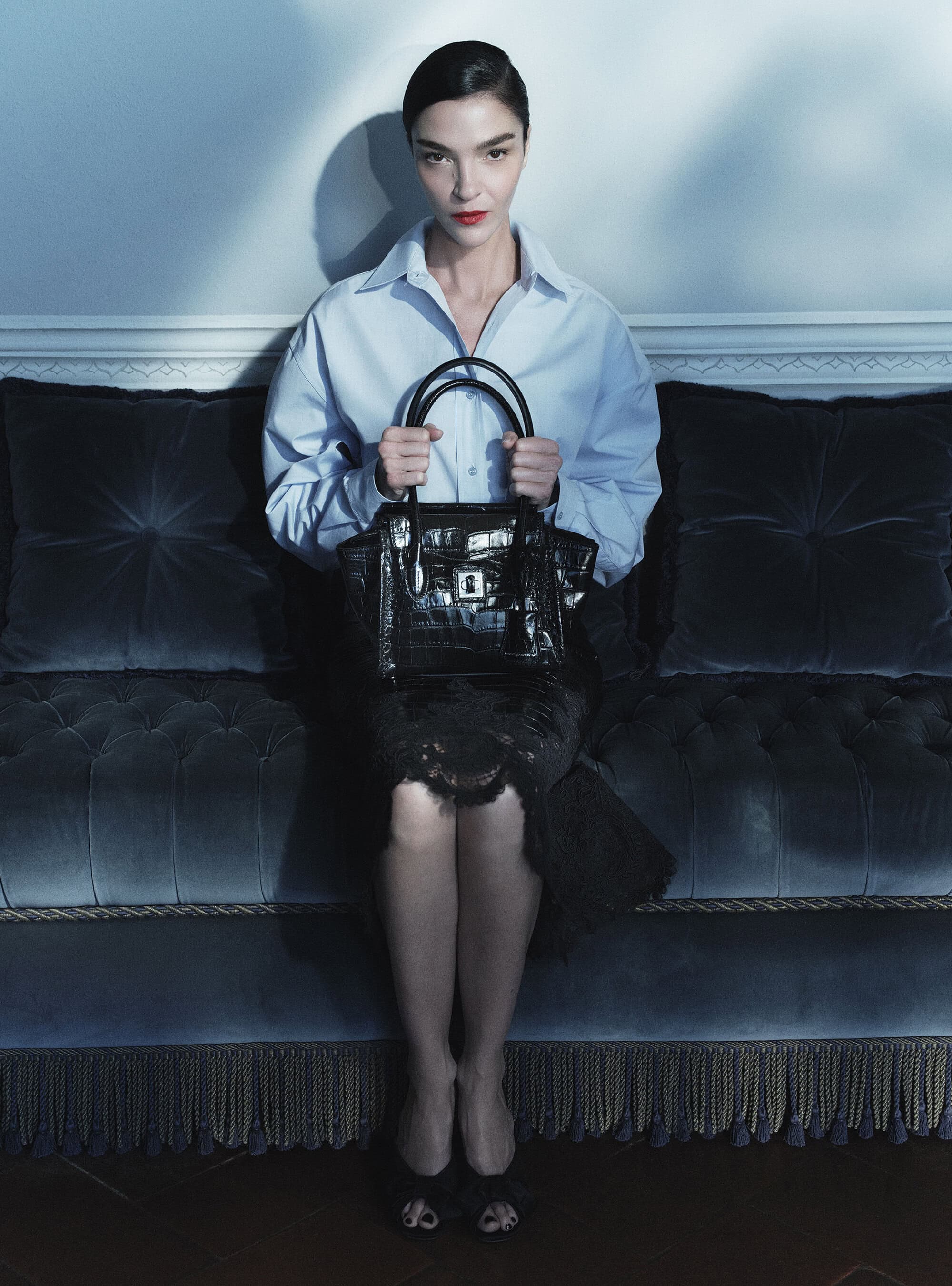 Mariacarla Boscono in a light blue shirt holding a black crocodile leather tote while seated on a tufted velvet sofa.
