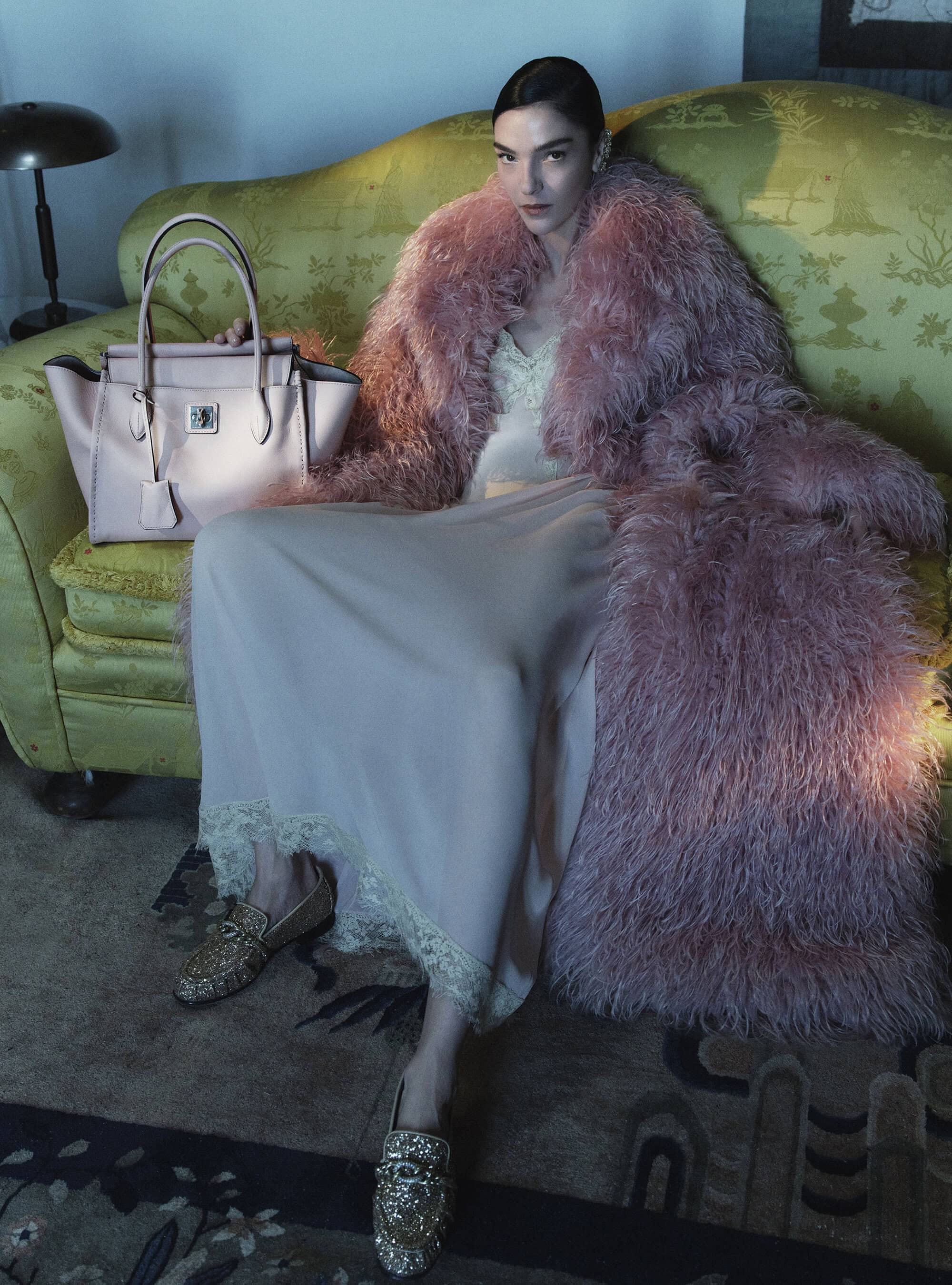 Model Mariacarla Boscono in a voluminous pink textured coat and pale slip dress seated on a green sofa beside a leather tote bag.