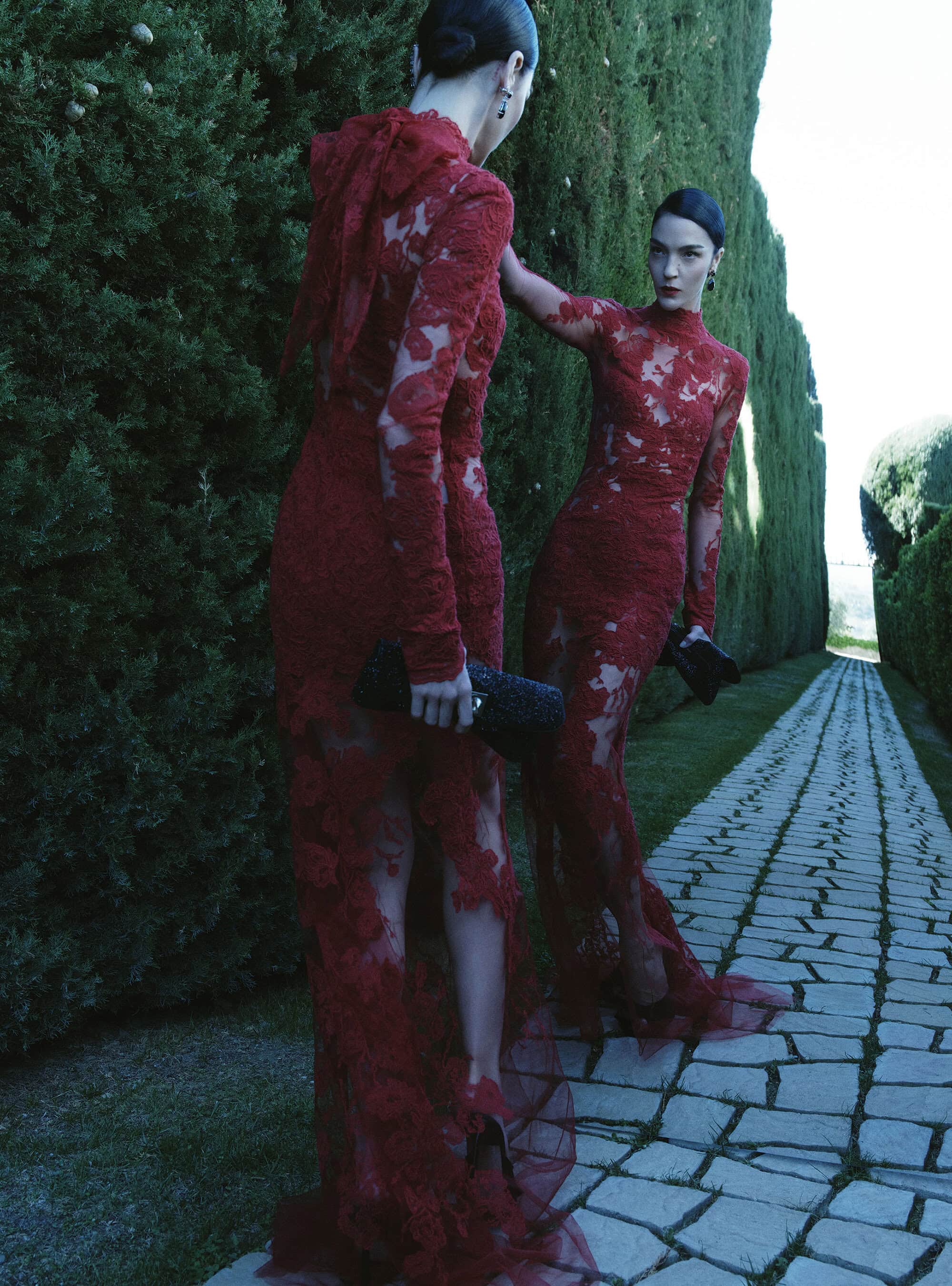 Mariacarla Boscono posing in a long-sleeved red lace gown beside a tall mirror reflection along a stone garden path.